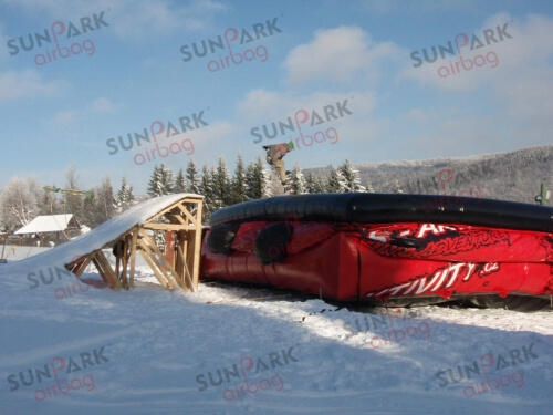 A red and black inflatable snowboarding airbag set up at the base of a snowy jump for snowboarders